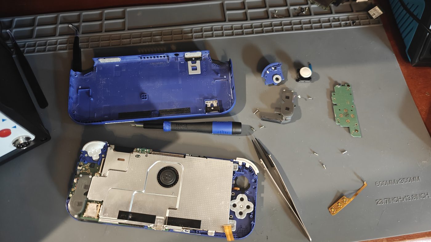 Nintendo Switch Lite opened on a repair mat — motherboard and blue housing separated, with the replacement right analog thumbstick module, a screwdriver and tweezers